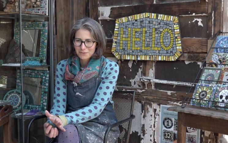 Mosaic artist Caroline Budden sits on a chair surrounded by examples of her mosaic work