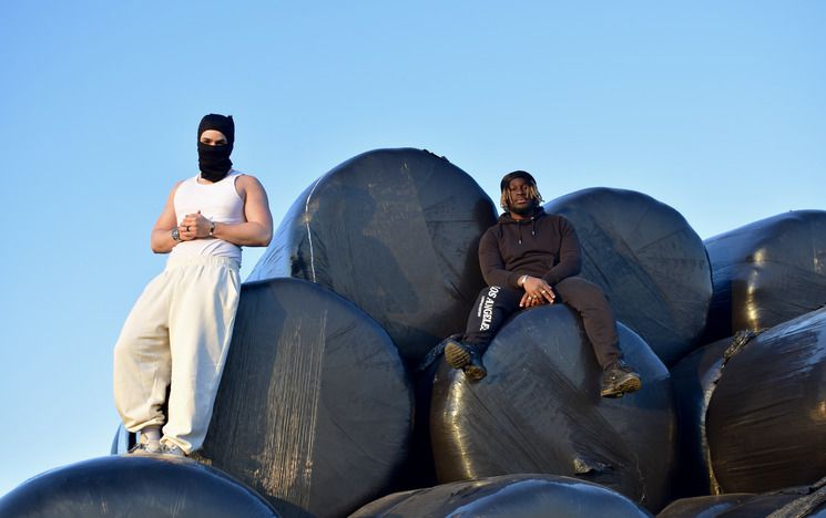 White man in white vest and jogging bottoms and black balaclava stands and Black man in black tracksuit sit on hay bales wrapped in black plastic