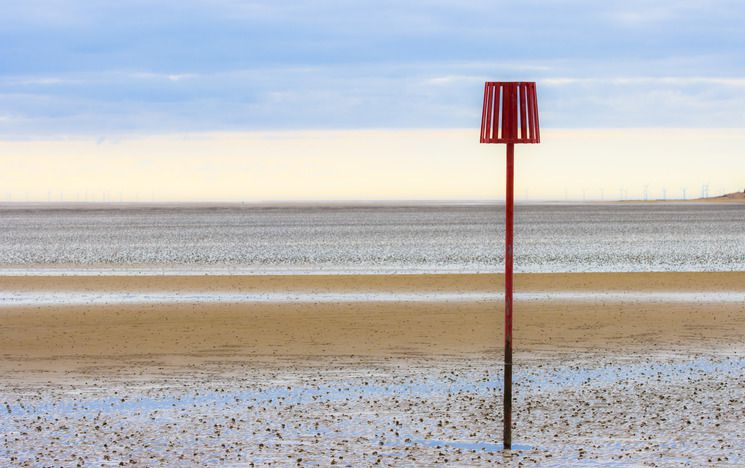 Beach scene at low tide showing sand. Red beach marker on right hand side of image. Wind turbines in the distance.