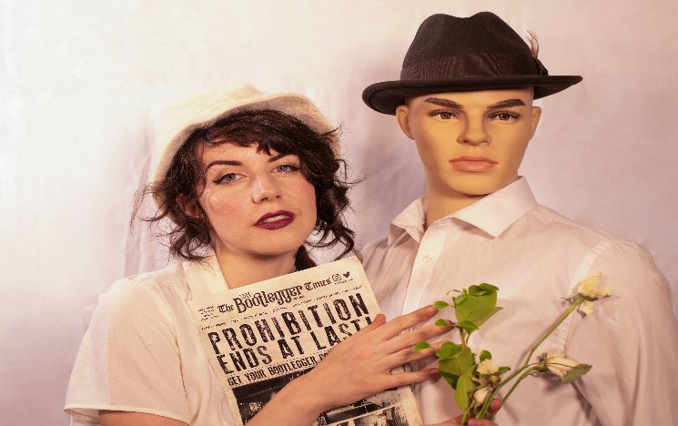 A woman is dressed as a bride, she is holding nearly dead flowers and a newspaper with headline about prohibition. She stands next to a male mannequin in a white shirt and trilby hat