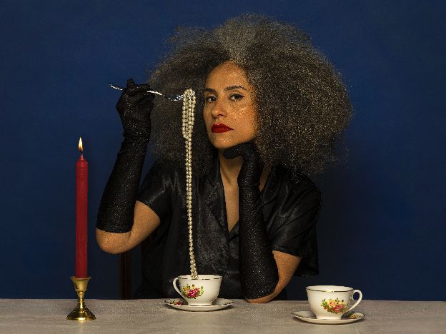 A black woman in black dress and black gloves sits at a table. The table has two cups and a lit red candle on it. She is holding a fork and pulling a pearl necklace out of the cup.