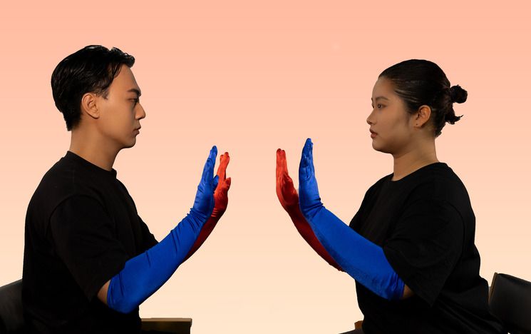 Two Asian people sit facing each other. They are wearing black clothes and have their hands up towards each other. They are wearing one red and blue glove each.