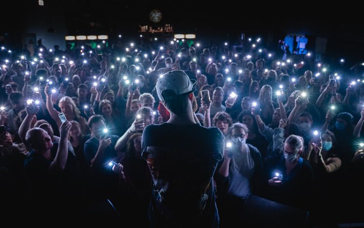 Back view of Musician Sam Tompkins looking out to audience. They are using torches on their phones and holding them up.