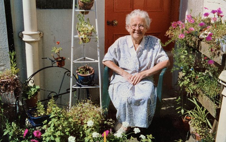 Old woman wearing a blue and white patterned dress sits on a green plastic chair in a small garden surrounded by flowers. She is smiling.