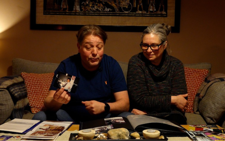 A masculine-presenting person and a feminine-presenting person browse through a photo album in their living room