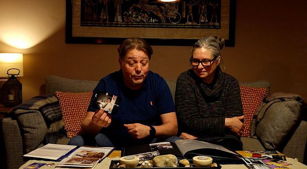 A masculine-presenting person and a feminine-presenting person browse through a photo album in their living room