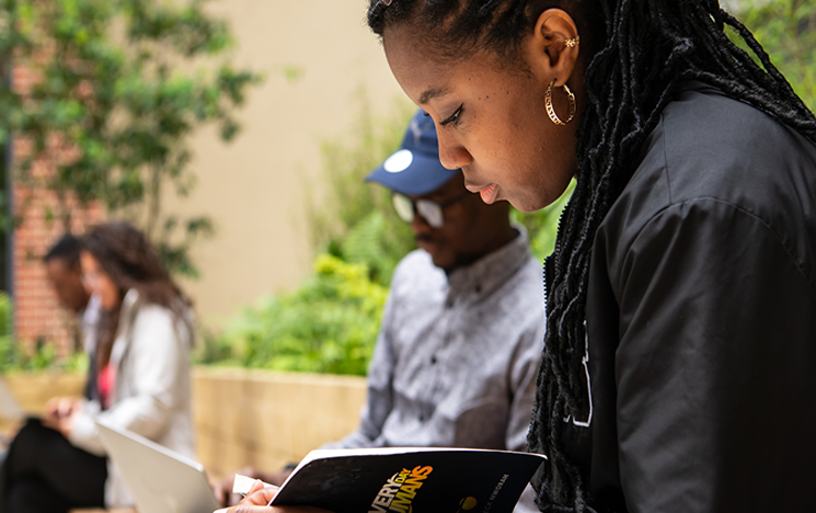 A Black femme presenting person reads a book in the foreground with other students in the background.