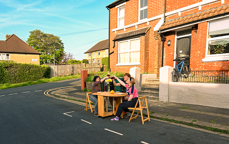 A group of people sitting round a wooden table on a street outside a house.