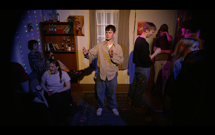 A man standing spotlighted inside a house as a party happens around him
