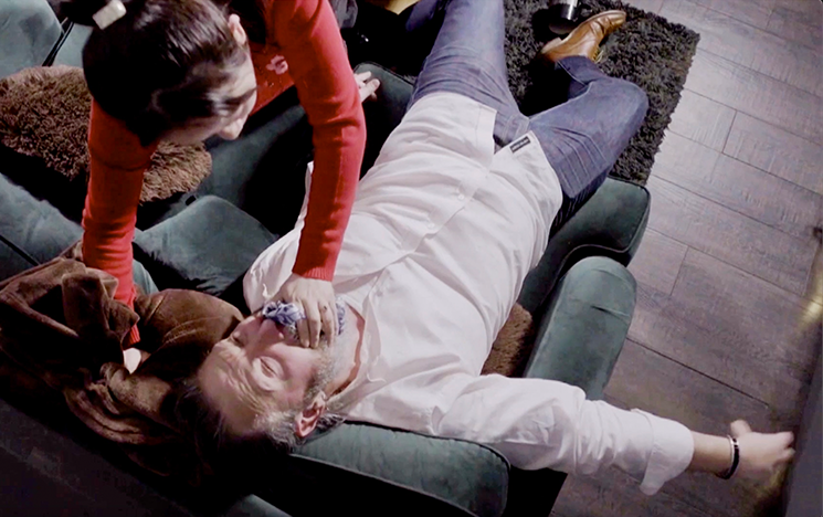 A woman uses a cloth to smother a man passed out on the sofa