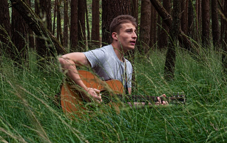 A man sitting in long grass playing a guitar.