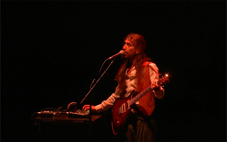 A man singing, playing guitar and reaching out for a keyboard