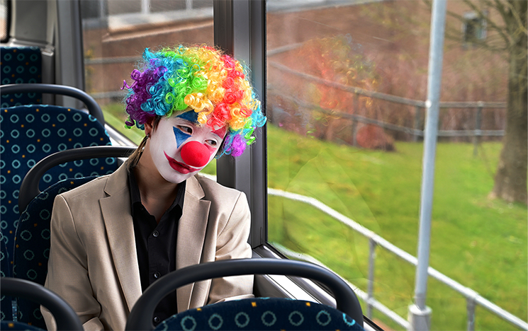 A woman with clown makeup and a rainbow wig on sitting on a bus looking out the window.