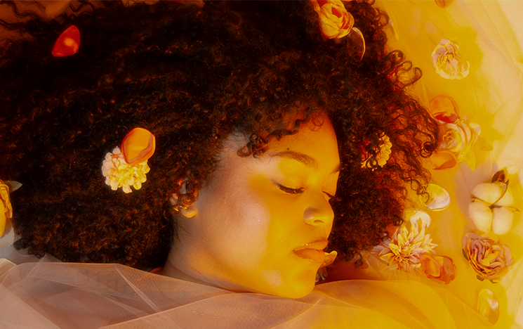 A woman lying down covered with a sheet, on a yellow background with flowers.
