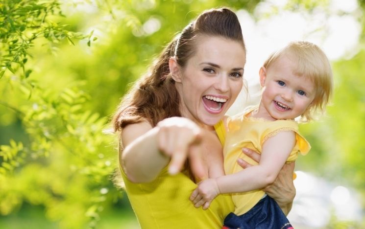 Smiling woman holds a toddler and points at the camera