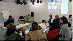 Actors sit around a table reading a scrupt, participants on a zoom call are projected onto the wall