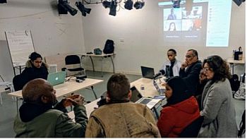 Actors sit around a table reading a scrupt, participants on a zoom call are projected onto the wall