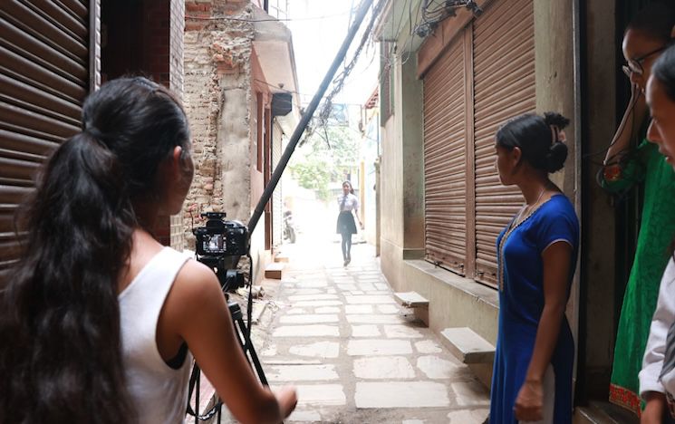 Camera rolling event image Photograph taken behind a teenage girl and a camera on a tripod in a residential alley. In the distance a girl walks towards the camera. On the right hand side, three girls stand on a doorstep watching the girl in the distance.