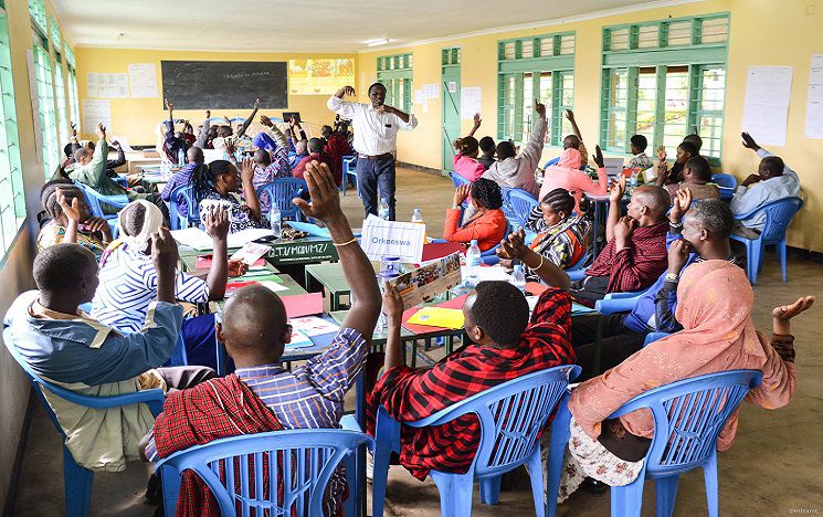 A group of people in a teaching room in Africa, raising their hands.