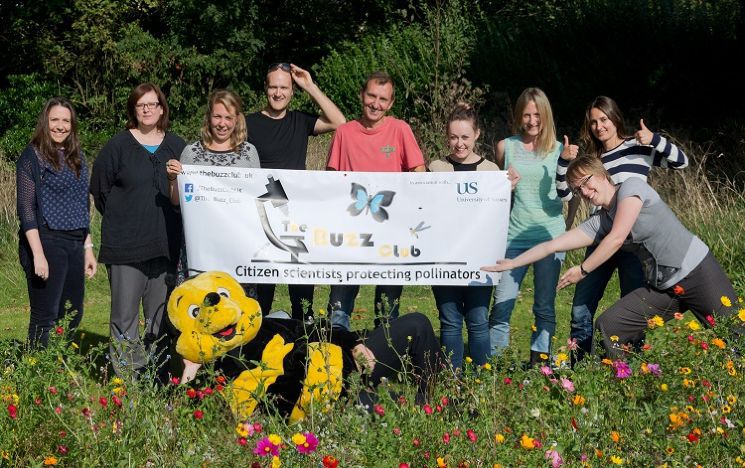 A group of people holding a sign saying 'the buzz club' in front of some wild flowers