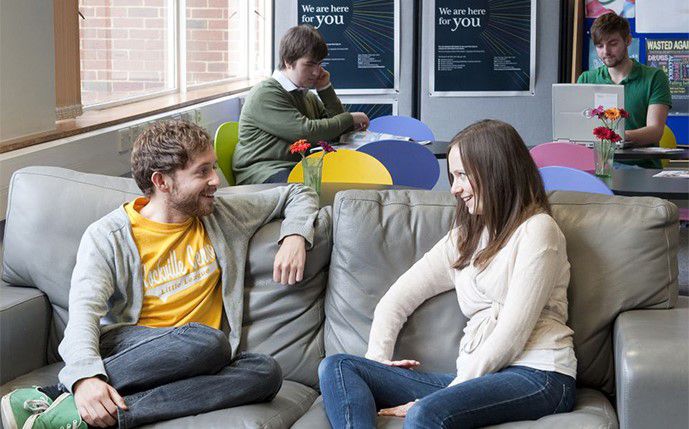 Two students talking on the sofa in the Student Life Centre, with Sussex signs in the background that read We are here for you