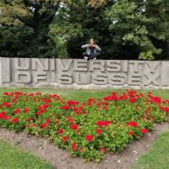 Aayushi, a graduate, kneeling on top of the Sussex Sign in front of a red heart made out of flowers