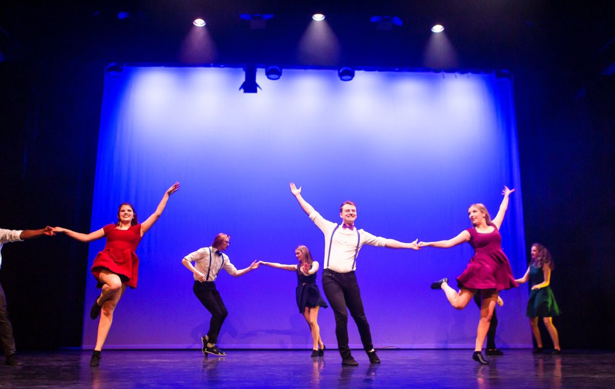 Several dancers in couples perform on a darkly lit stage