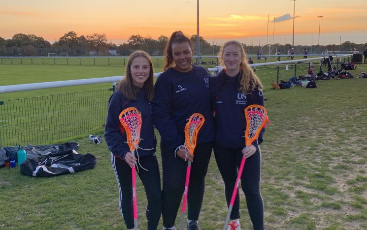 Three Lacrosse students posing for a photo infront of a sunset on a Lacrosse pitch