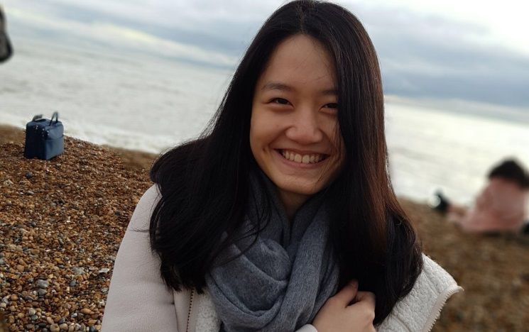 Student smiling close up on pebbly beach with sea behind