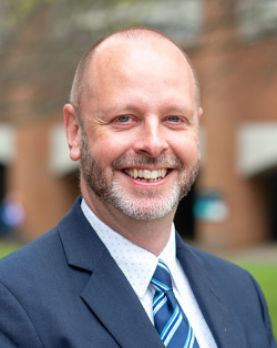 Man in a suit poses for a photo smiling towards camera, campus in background