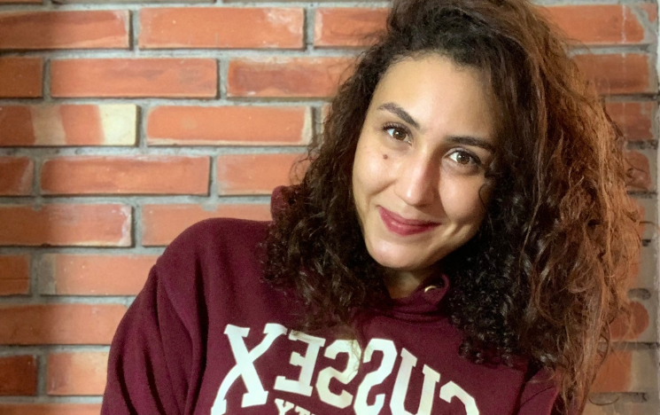 Photo of Soumia Chajai. She is smiling at the camera and is on the Sussex campus, wearing a maroon Sussex jumper