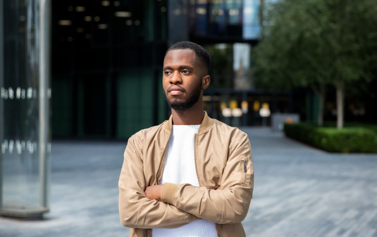 A headshot photo of alumnus Wilfrid Obeng. He has his arms crossed in a tan jacket and white tshirt