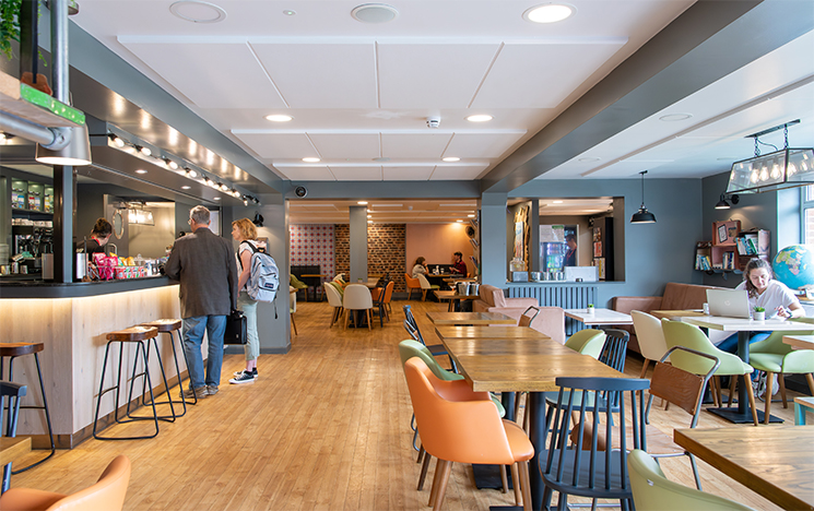 Interior of IDS bar and kitchen, showing tables and chairs to the right of the picture, and a bar on the left, with people ordering food from a member of staff