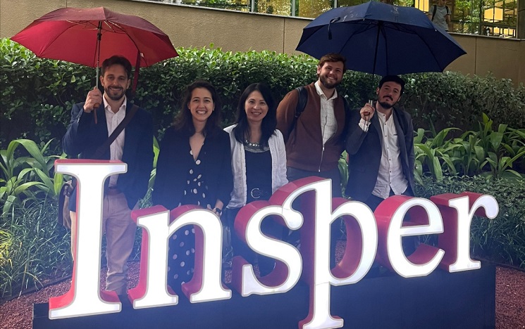 People standing under umbrellas in front of a glowing sign for INSPER