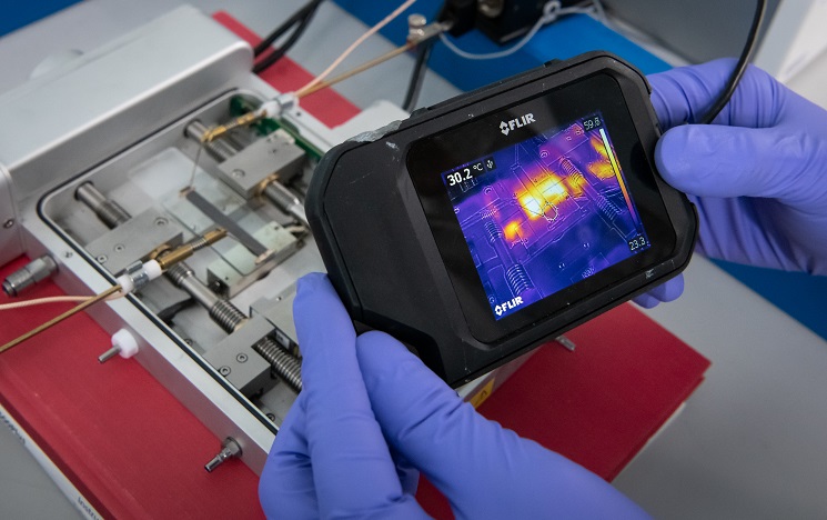 A closeup of a pair of blue-gloved hands looking at a machine through a thermal camera, with a digital image of warmer and cooler areas displayed on the camera screen