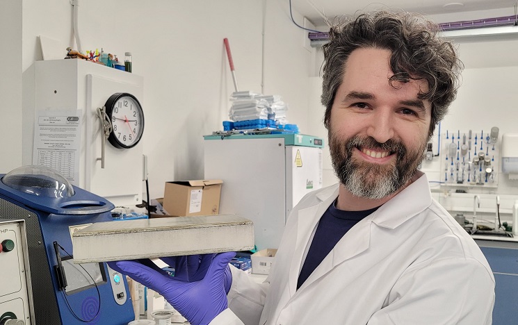 A white-coated scientist in a lab smiling as he holds up a cross section of a wind turbine blade in his gloved hands