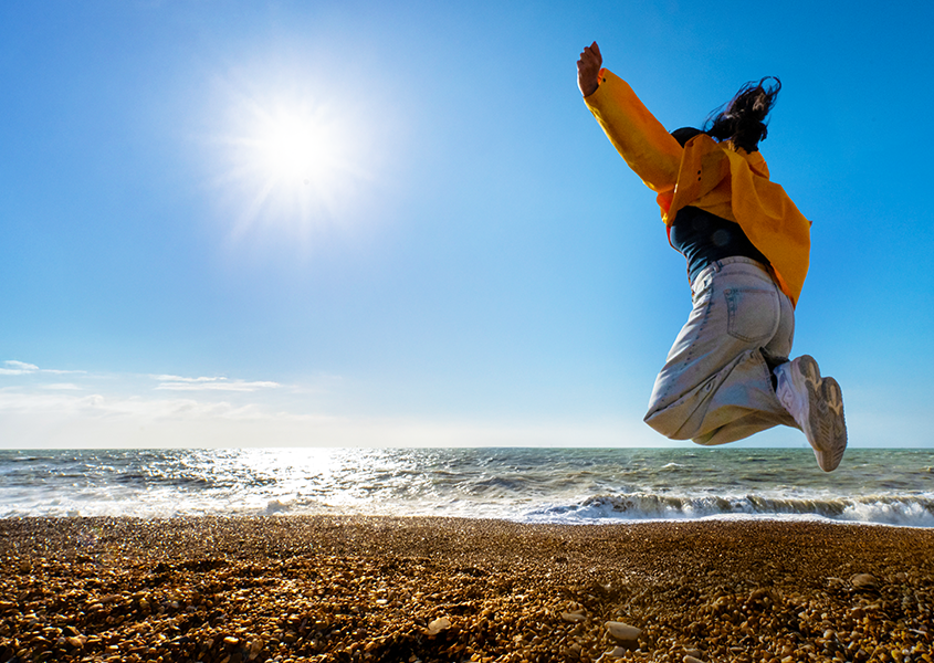 Person in orange jacket jumping enthusiastically on sunny Brighton beach