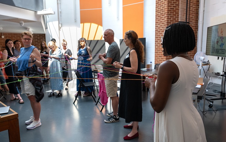A group of people standing in a sunlit room connected by lengths of coloured thread