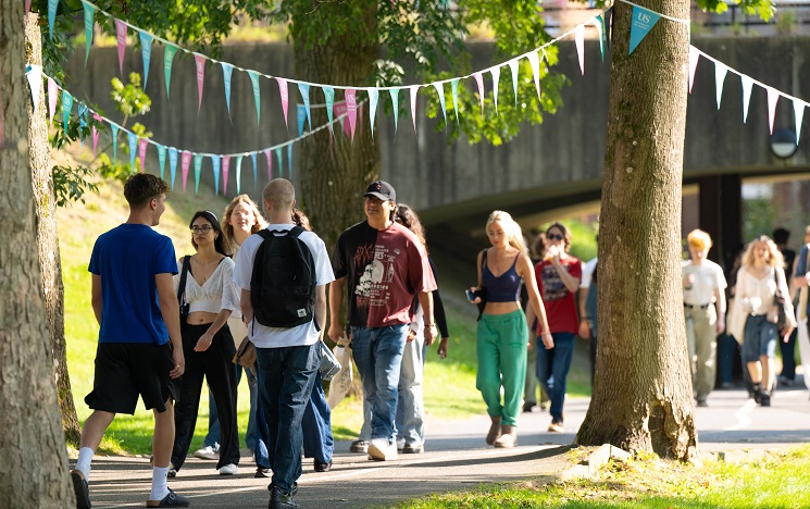 Students walking along a sunny tree-lined avenue on the University of Sussex campus, underneath strings of colourful bunting