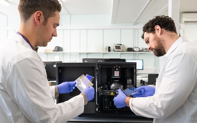 Two scientists wearing white coats and blue latex gloves looking at objects through plastic spectroscopy equipment