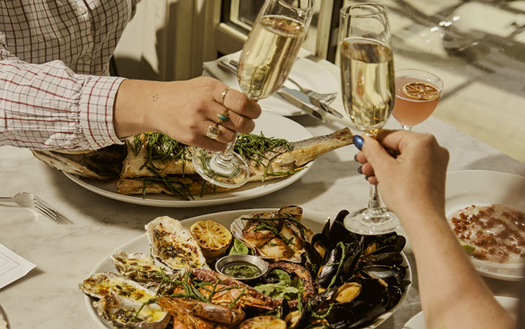 Two people toasting champagne with a seafood platter at a dinner table.