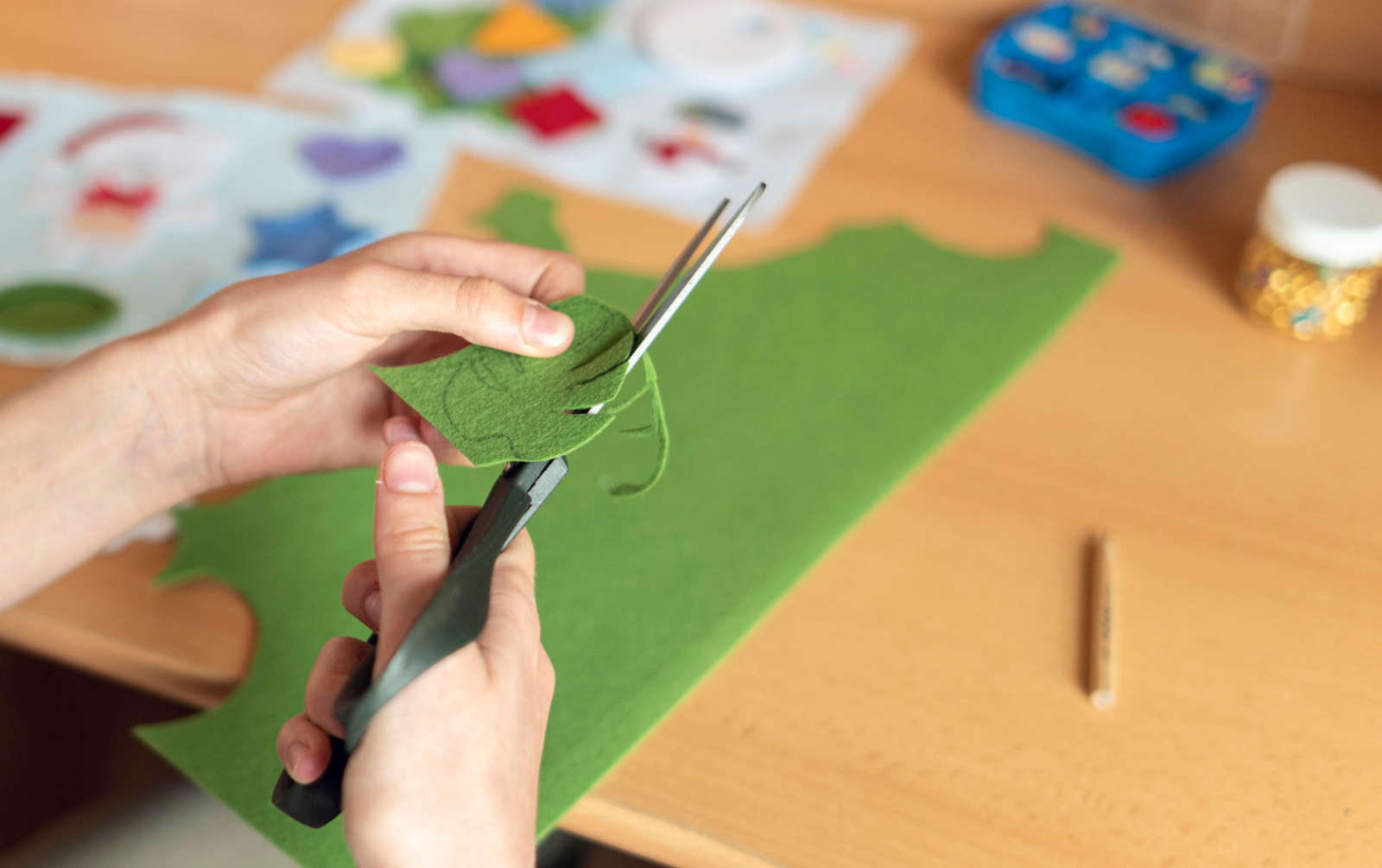Hands cutting a green felt leaf with scissors on a table. Craft materials like paper and coloured boxes are visible, conveying a creative, focused atmosphere.