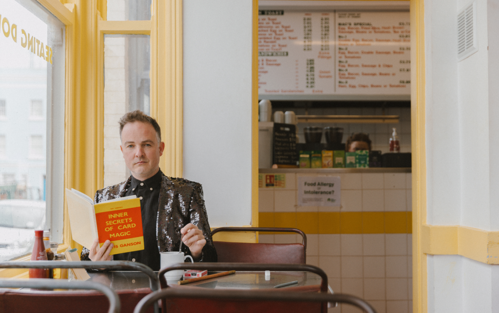 Augusto Corrieri in a sequin jacket reads 'Inner Secrets of Card Magic' at a cafe. The menu on the wall and condiments on the table create a casual, curious vibe.