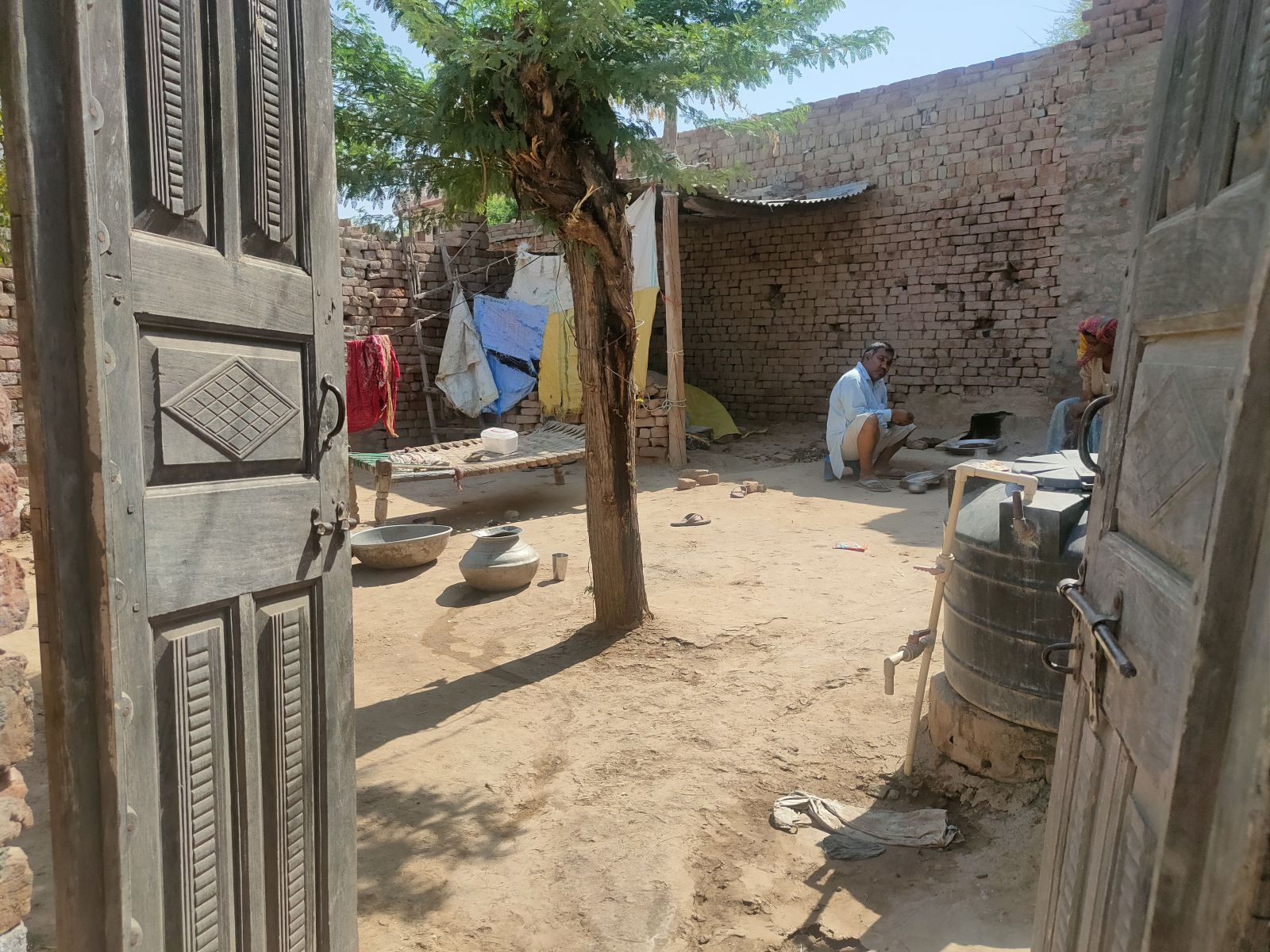 View of a courtyard, there is a man with some pots, a washing line with old looking cloth on it, other various items are across the courtyard. 