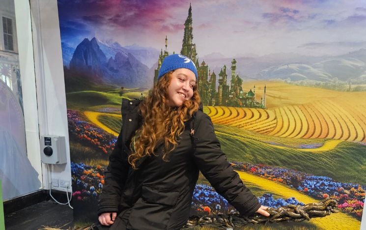 Alumna Megan Bond sits in front of a large, colourful backdrop depicting a fantasy landscape with fields, mountains and a castle.