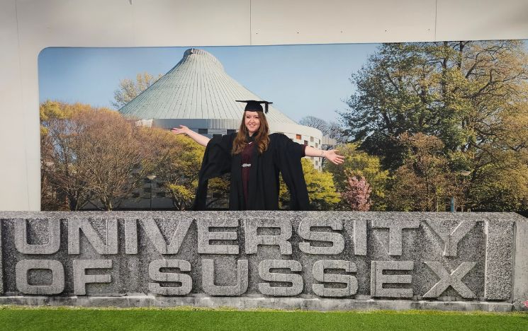Alumna Megan Bond at graduation standing behind the &lsquo;University of Sussex&rsquo; graduation sign wearing her graduation gown. In the background, the Meeting House and trees.