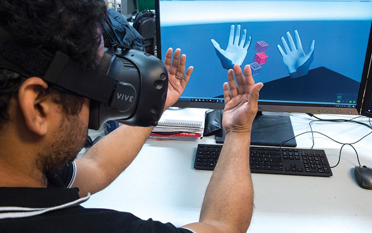A student in a headset raising his hands while they display in virtual reality on a computer screen