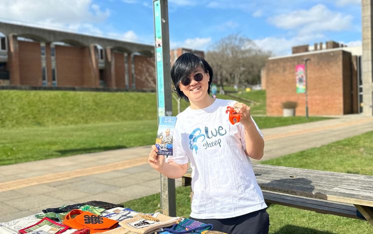 Alumna Kacci Xia behind a stall displaying craft goods in Library Square on campus. Kacci is holding a leaflet and a key chain.