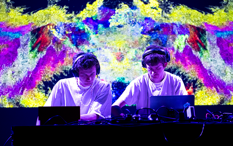 Two students perform a DJ set in front of a colourful background screen.