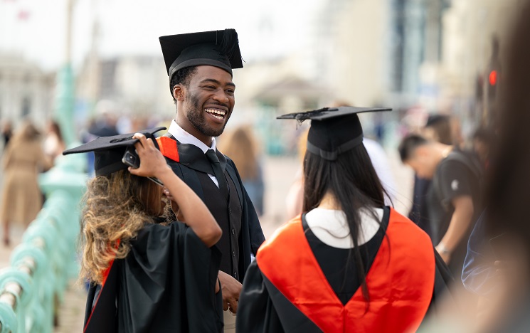 Three graduates wearing mortarboard hats and black academic robes with red hoods, laughing on Brighton seafront